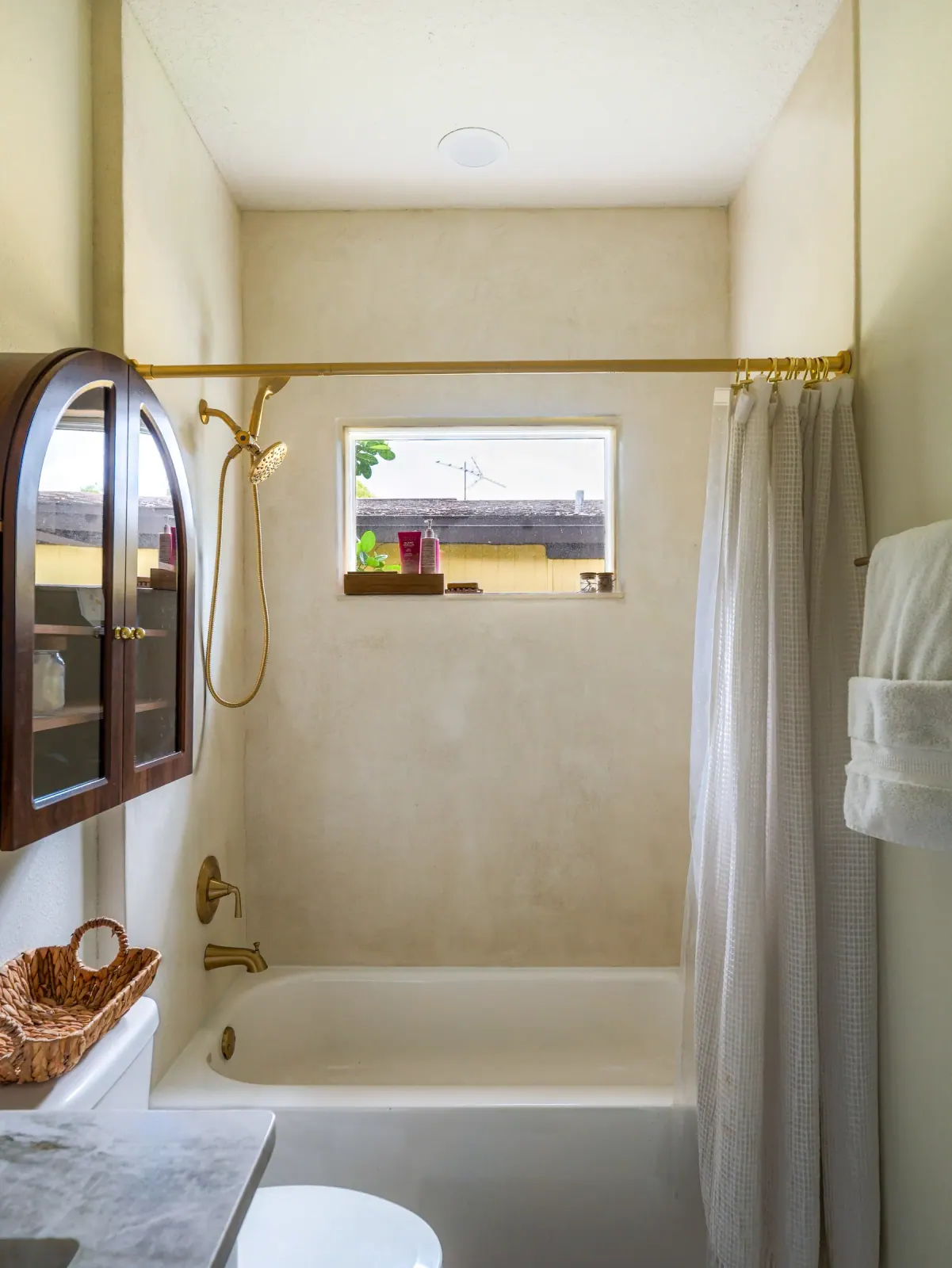 Microcement tub-and-shower surround with brass fixtures and a walnut arched cabinet